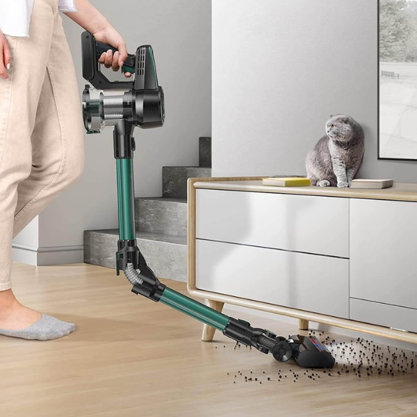 Woman using a green cordless stick vacuum on a wood floor while a cat sits on a cabinet