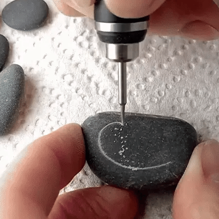 Hand using a cordless engraving pen to carve a curved line into a dark pebble